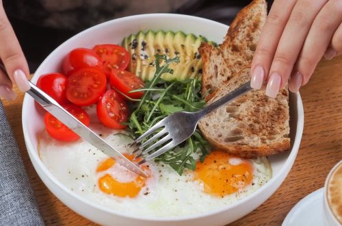 Close-up of a breakfast bowl with fried eggs, avocado, tomatoes, and bread, served with cappuccino.
