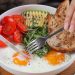 Close-up of a breakfast bowl with fried eggs, avocado, tomatoes, and bread, served with cappuccino.